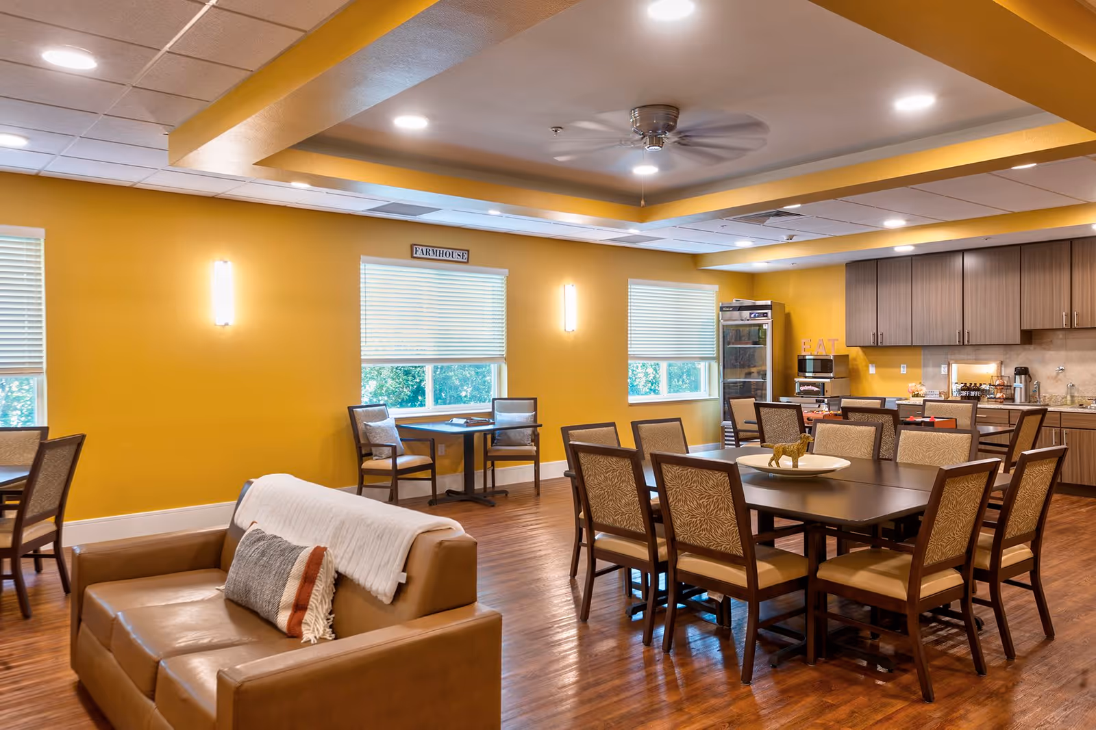 Bright communal dining and lounge area with tables and chairs, a kitchen counter, and a sofa against yellow walls.