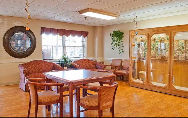 A cozy common area with a wooden table surrounded by four chairs, two patterned sofas, and two additional chairs against the wall. A large round clock hangs on the wall near a window with blinds and a valance. There is a wooden display cabinet with glass doors on the right side and a hanging plant on the wall. The room has wooden flooring and ceiling lights.