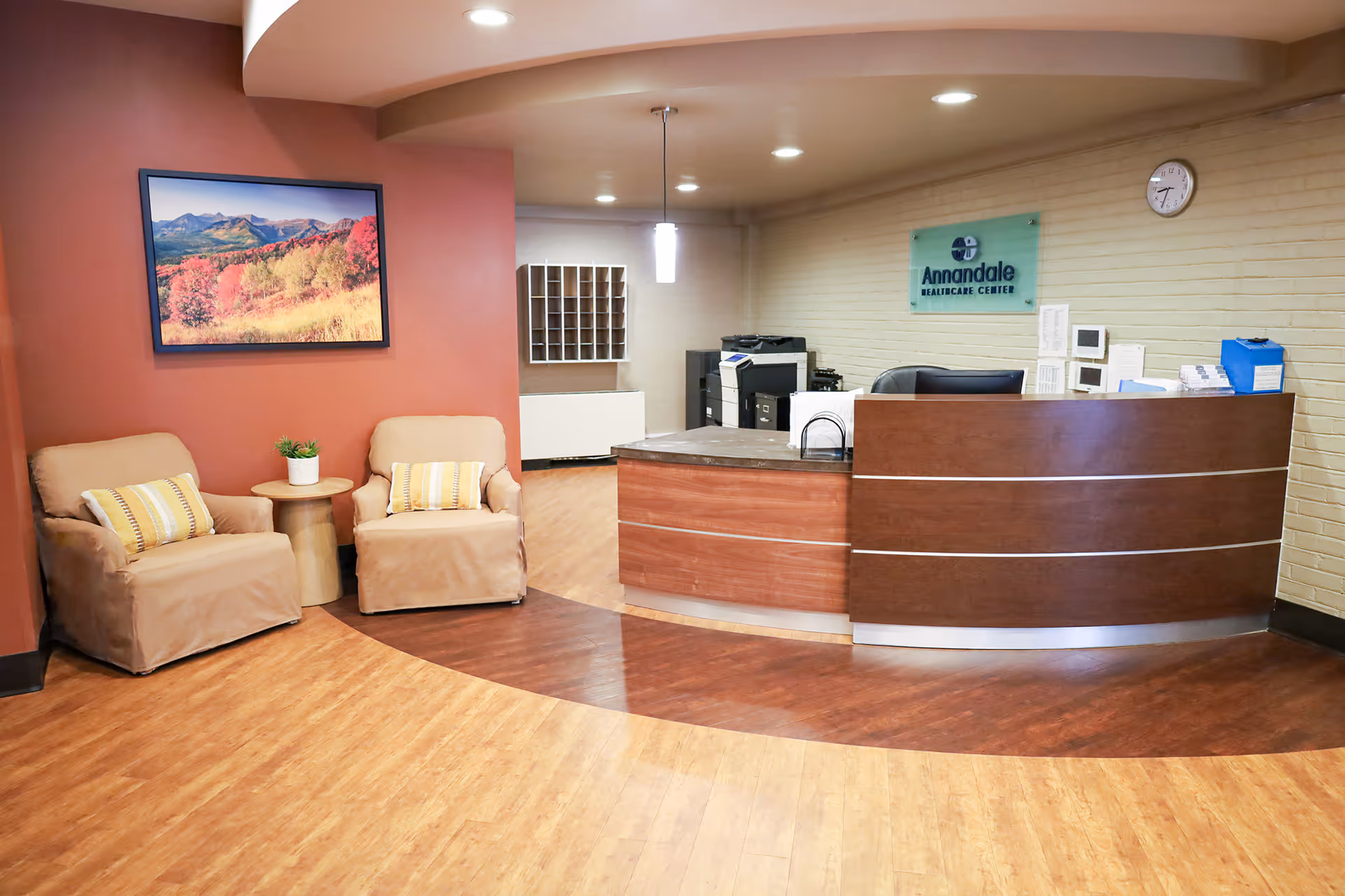 Reception area of Annandale Healthcare Center featuring a curved wooden reception desk with office equipment behind it, two beige armchairs with striped pillows and a small round table with a plant, a large landscape painting on a red accent wall, and a clock on a beige brick wall.