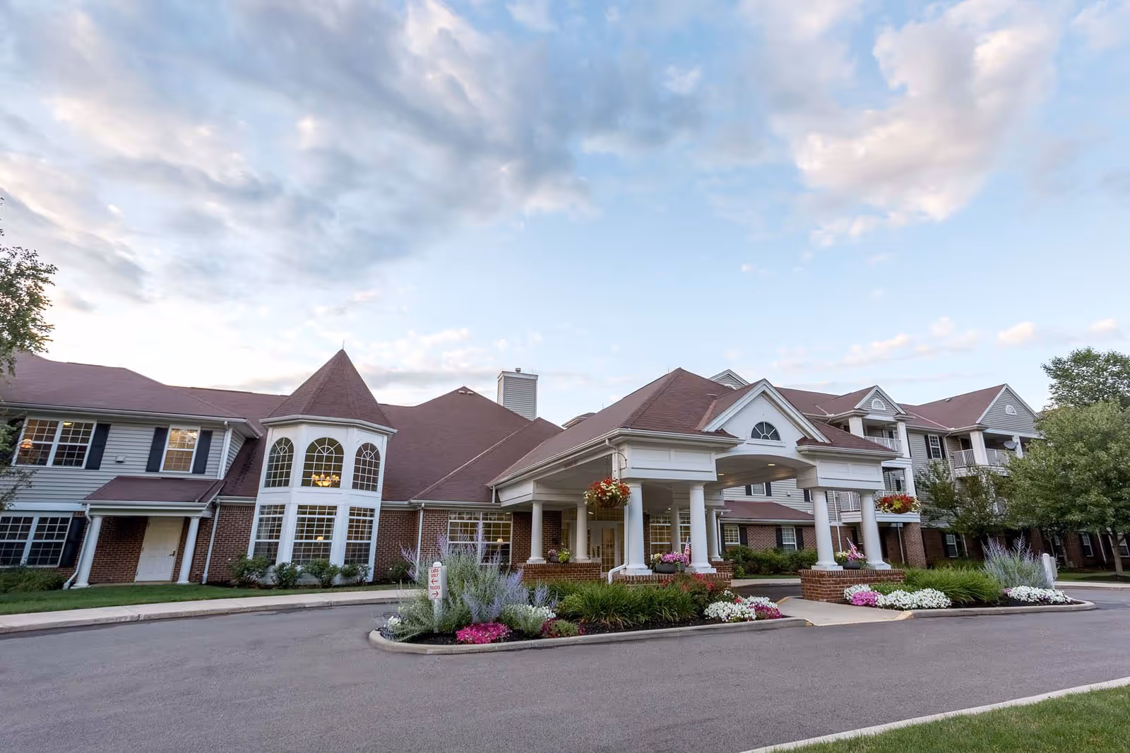 Exterior view of Independence Village of Avon Lake, showing a large building with a covered entrance supported by white columns, red brick and white siding walls, multiple windows, and a landscaped area with flowers and greenery in front. The sky is partly cloudy.
