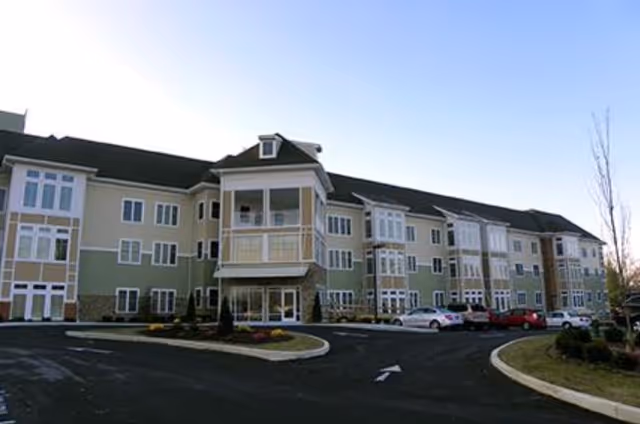 Exterior view of a multi-story senior living facility building with multiple windows, a covered balcony, and a parking lot with several cars parked. The sky is clear and the surrounding area has some landscaping with small trees and bushes.