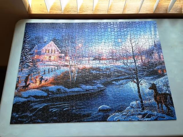 A completed jigsaw puzzle of a snowy winter cabin and river scene with a deer, displayed on a table beneath window blinds.