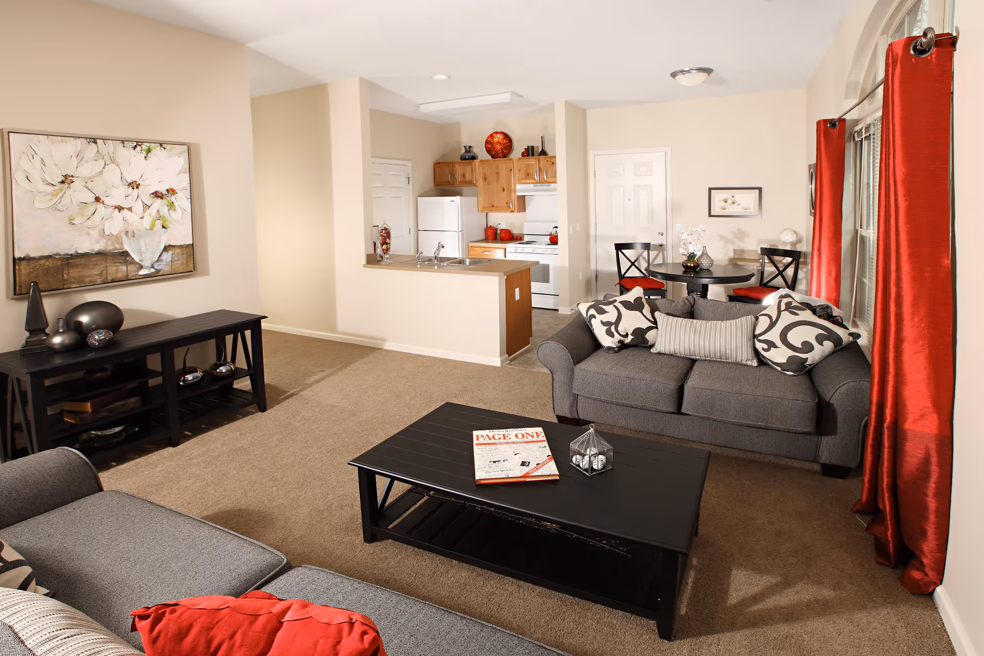 A cozy living room area with two gray sofas adorned with patterned and striped pillows, a black coffee table with a magazine and decorative item, and a black console table with decorative objects. The room has beige walls, carpeted floor, red curtains on a large window, and a round dining table with four chairs in the background near a small kitchen with wooden cabinets, white appliances, and a countertop.