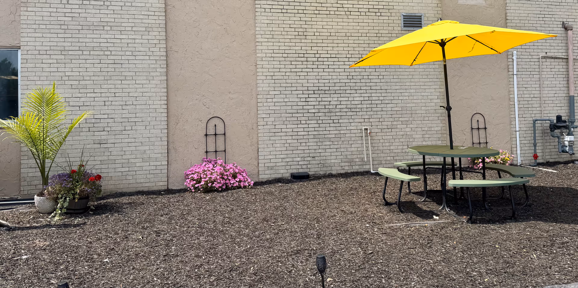 Outdoor area with a round green picnic table and attached benches under a large yellow umbrella. The ground is covered with mulch, and there are potted plants and flowers near a beige brick wall in the background.