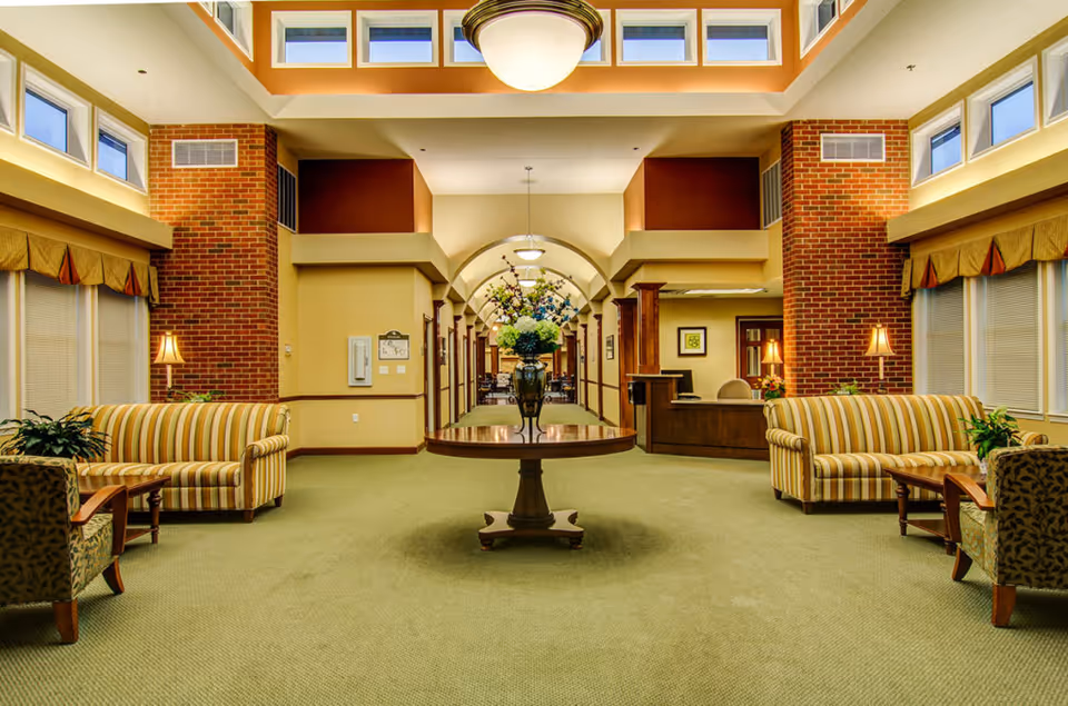 A spacious and well-lit common area with green carpet, two striped sofas facing each other, two armchairs, a round wooden table with a large floral arrangement in the center, brick pillars, multiple windows near the ceiling, and a reception desk in the background.