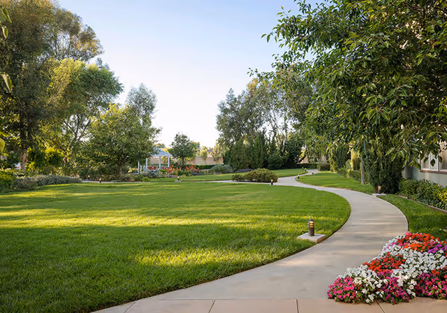 Curving paved walkway through a well-kept lawn and landscaped garden with trees and flowerbeds.