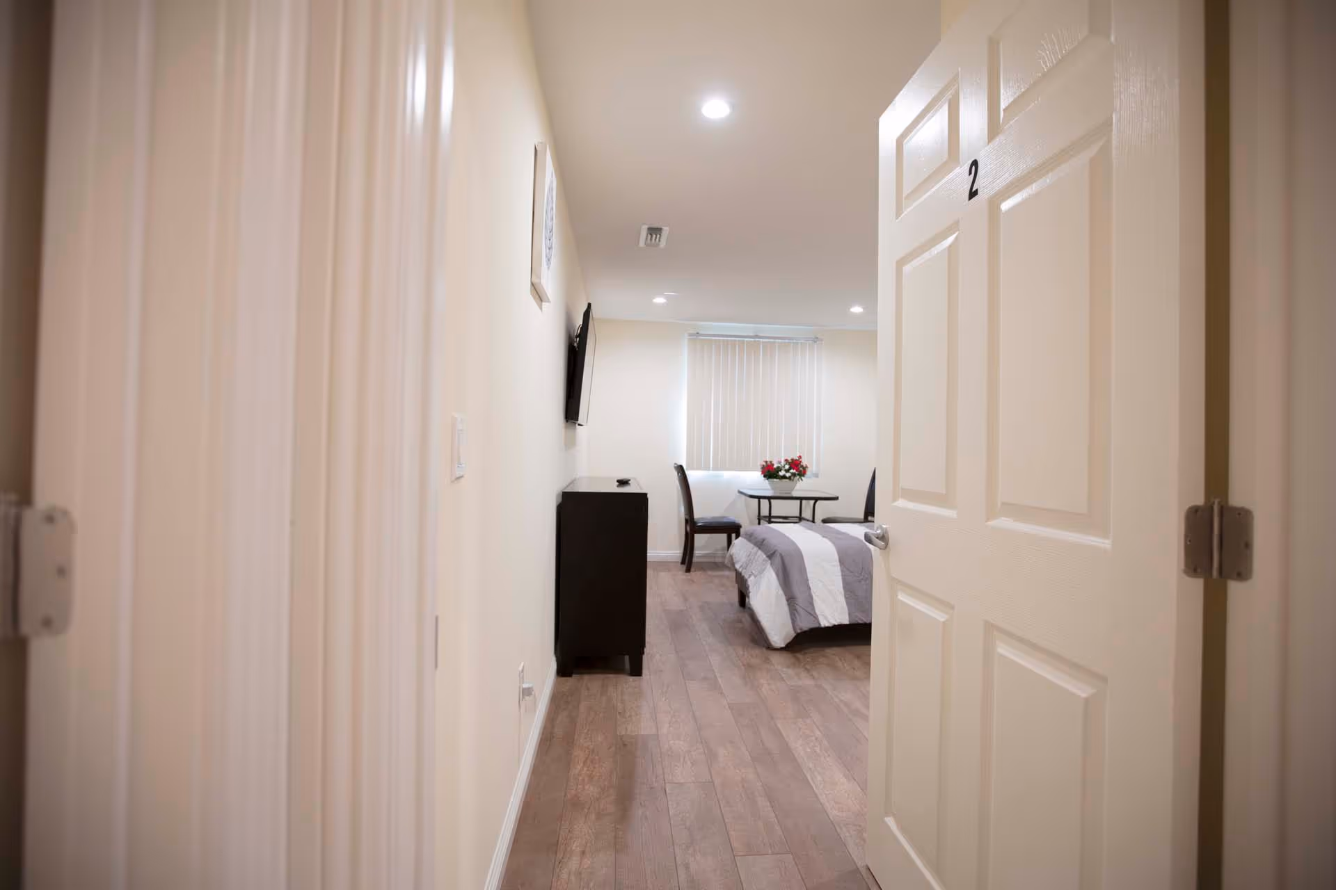 View through an open door labeled number 2 into a small bedroom with a bed covered in a gray and white striped comforter, a small table with two chairs, a vase of flowers on the table, a wall-mounted TV, and a dresser. The room has wood flooring and a window with vertical blinds.