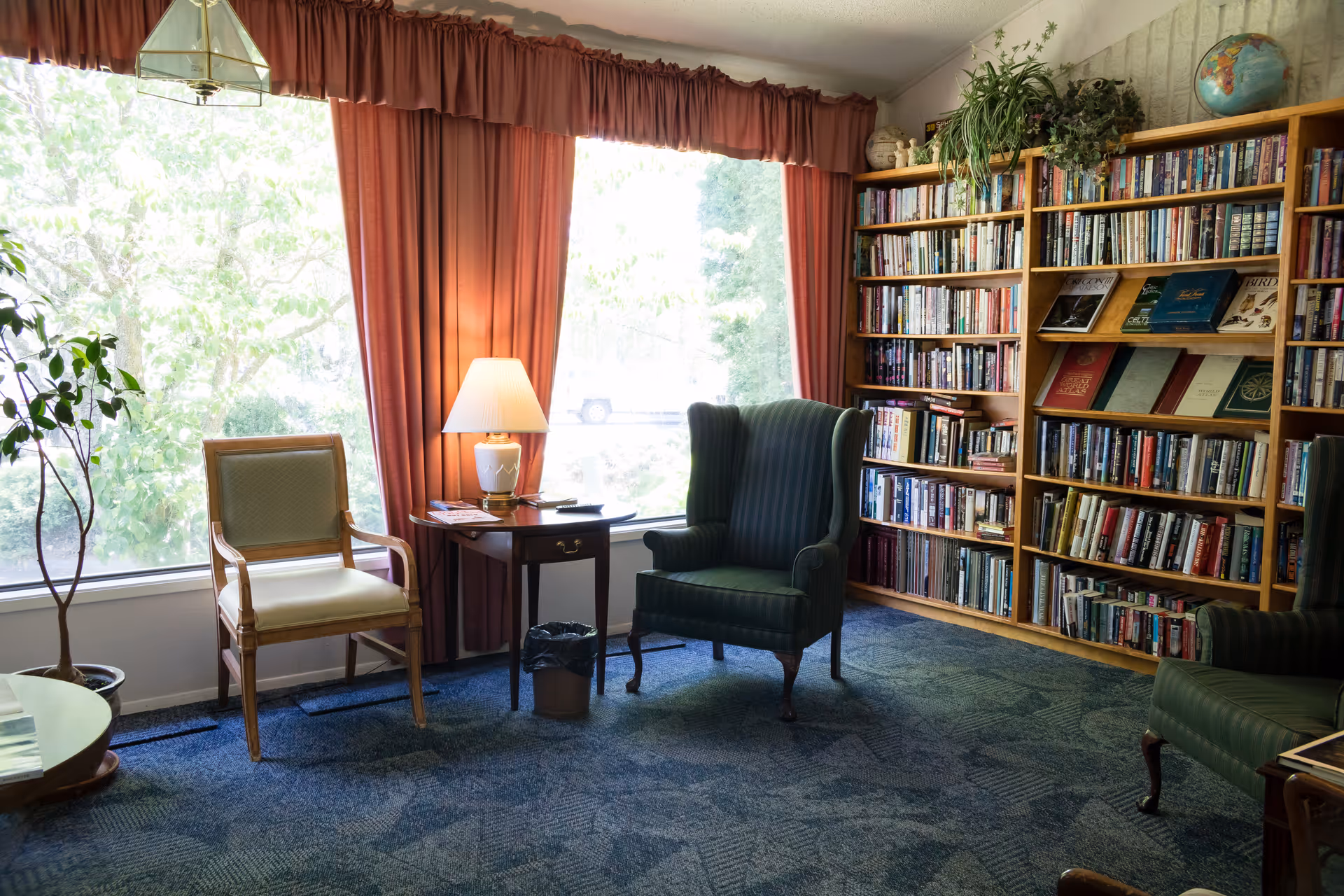 A cozy reading room with large windows covered by red curtains, a wooden bookshelf filled with books, two green upholstered armchairs, a wooden chair with a cushion, a small wooden side table with a lamp, and a potted plant near the window.