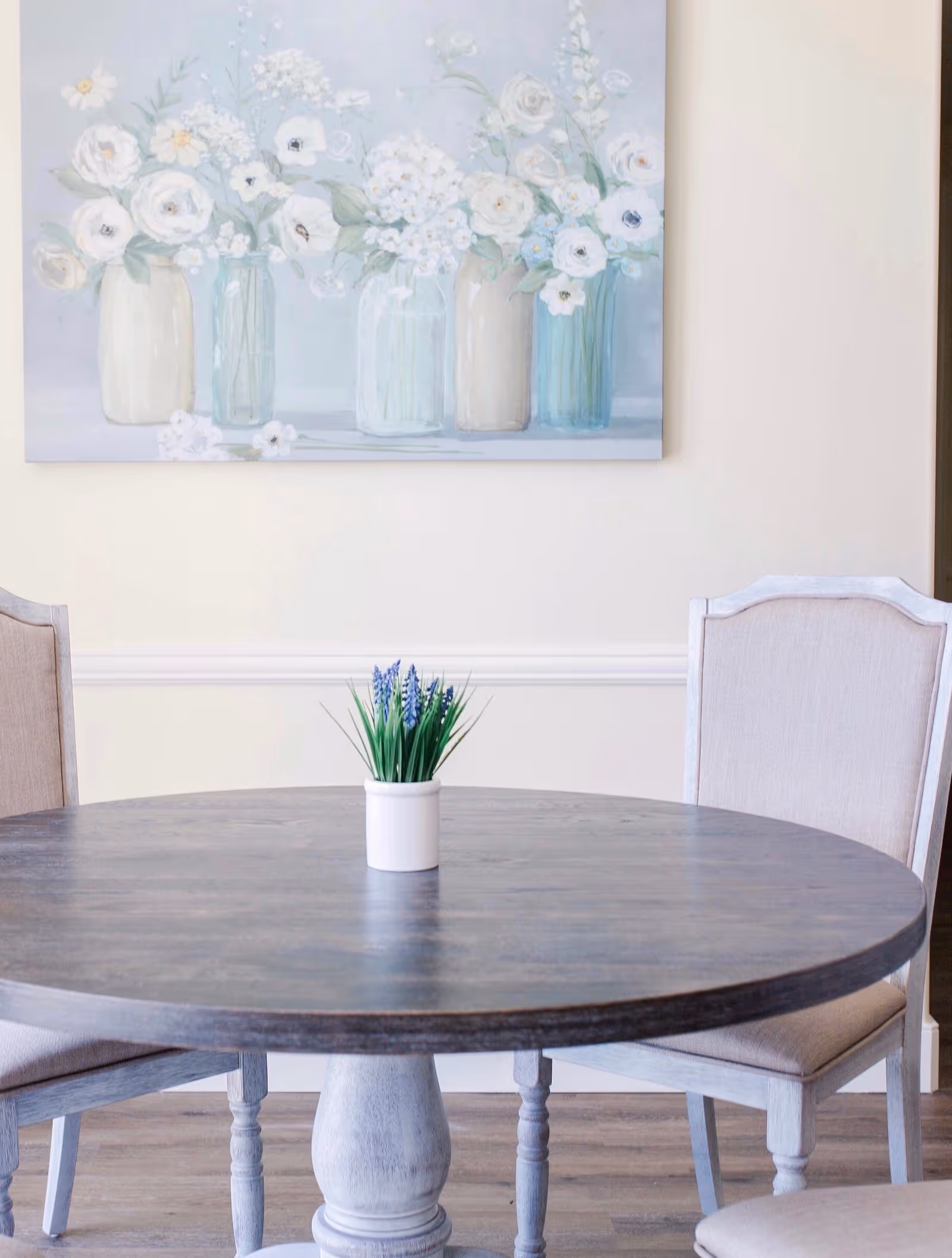 A round wooden dining table with a small white pot containing green plants and purple flowers in the center. Two upholstered chairs with light-colored fabric are positioned around the table. A painting of white flowers in vases hangs on the light-colored wall behind the table.