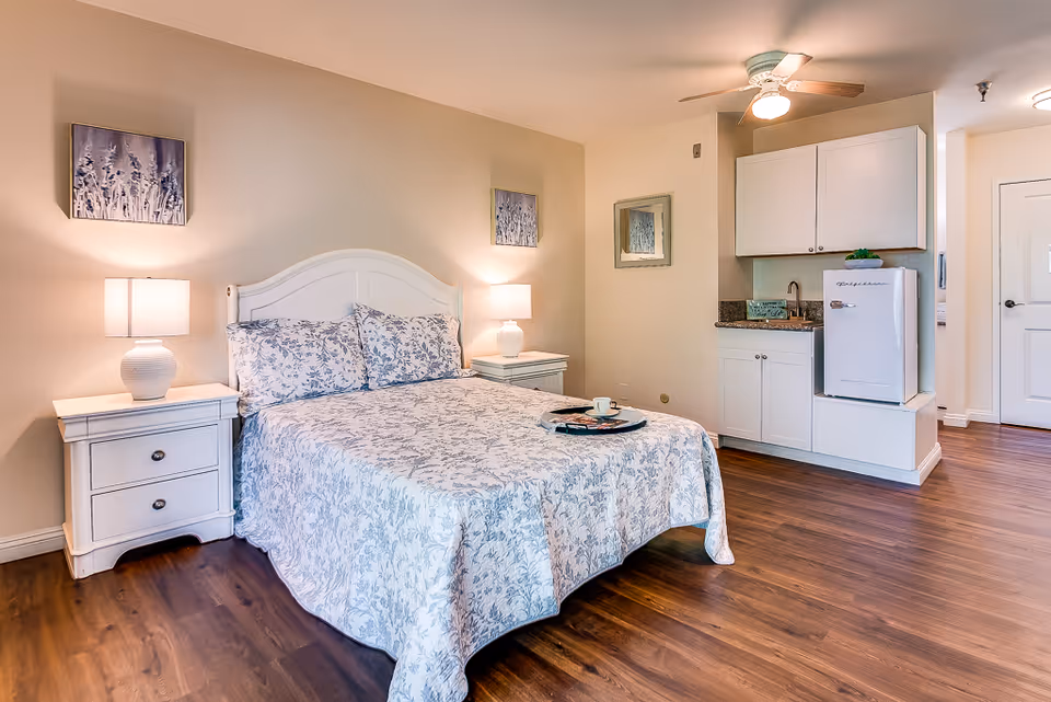A cozy senior living bedroom with a white bed frame and floral bedding. There are two matching nightstands with lamps on either side of the bed. The room has wooden flooring and light-colored walls with two framed artworks above the bed. To the right, there is a small kitchenette area with white cabinets, a mini refrigerator, a sink, and a ceiling fan above.