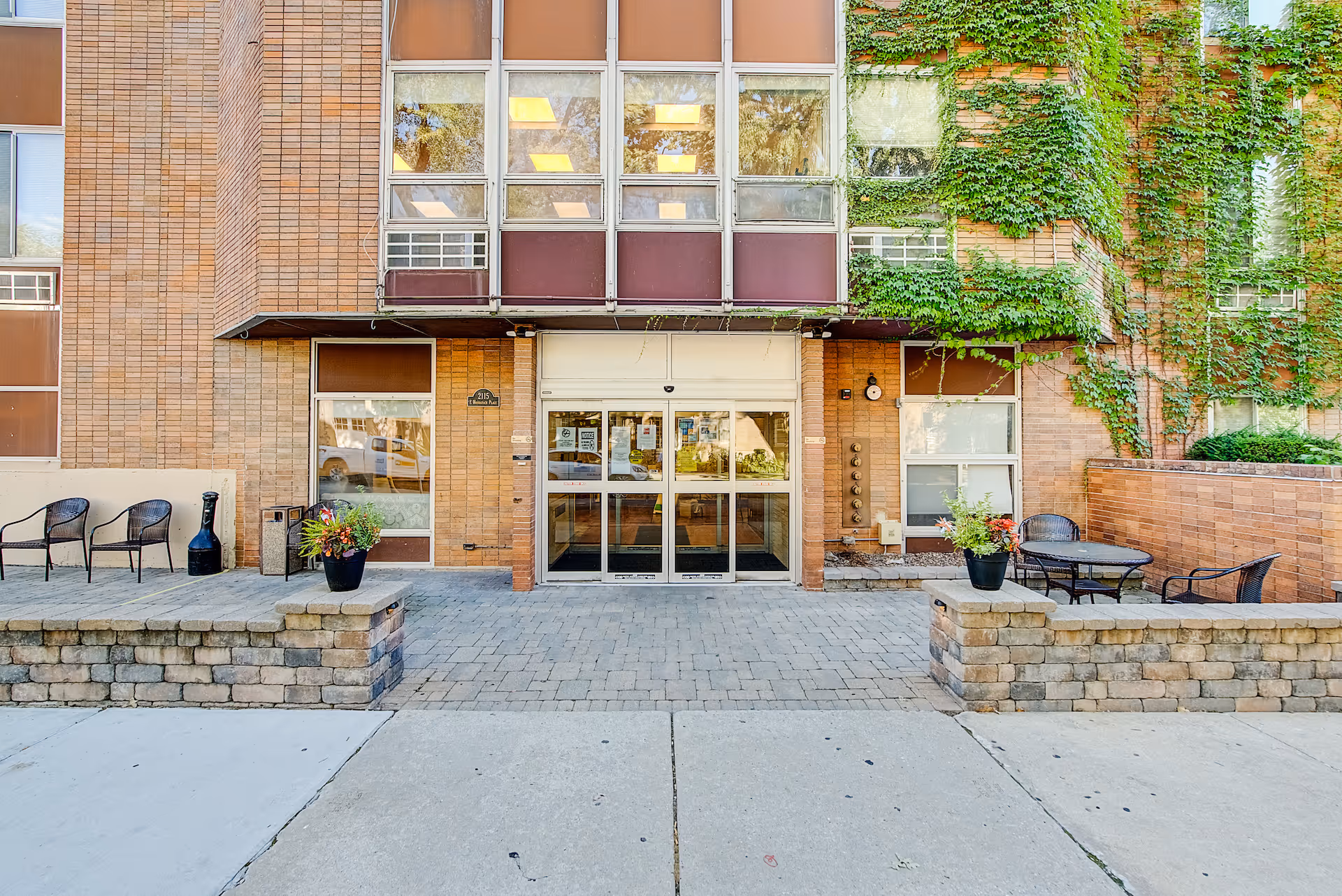 Entrance of a brick building with large glass doors, surrounded by windows and ivy growing on the walls. There are two small patio areas on either side of the entrance with black chairs, tables, and potted plants.