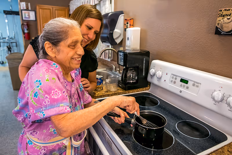 An elderly woman stirs a pot on an electric stovetop while a smiling caregiver stands behind her in a small kitchen area.