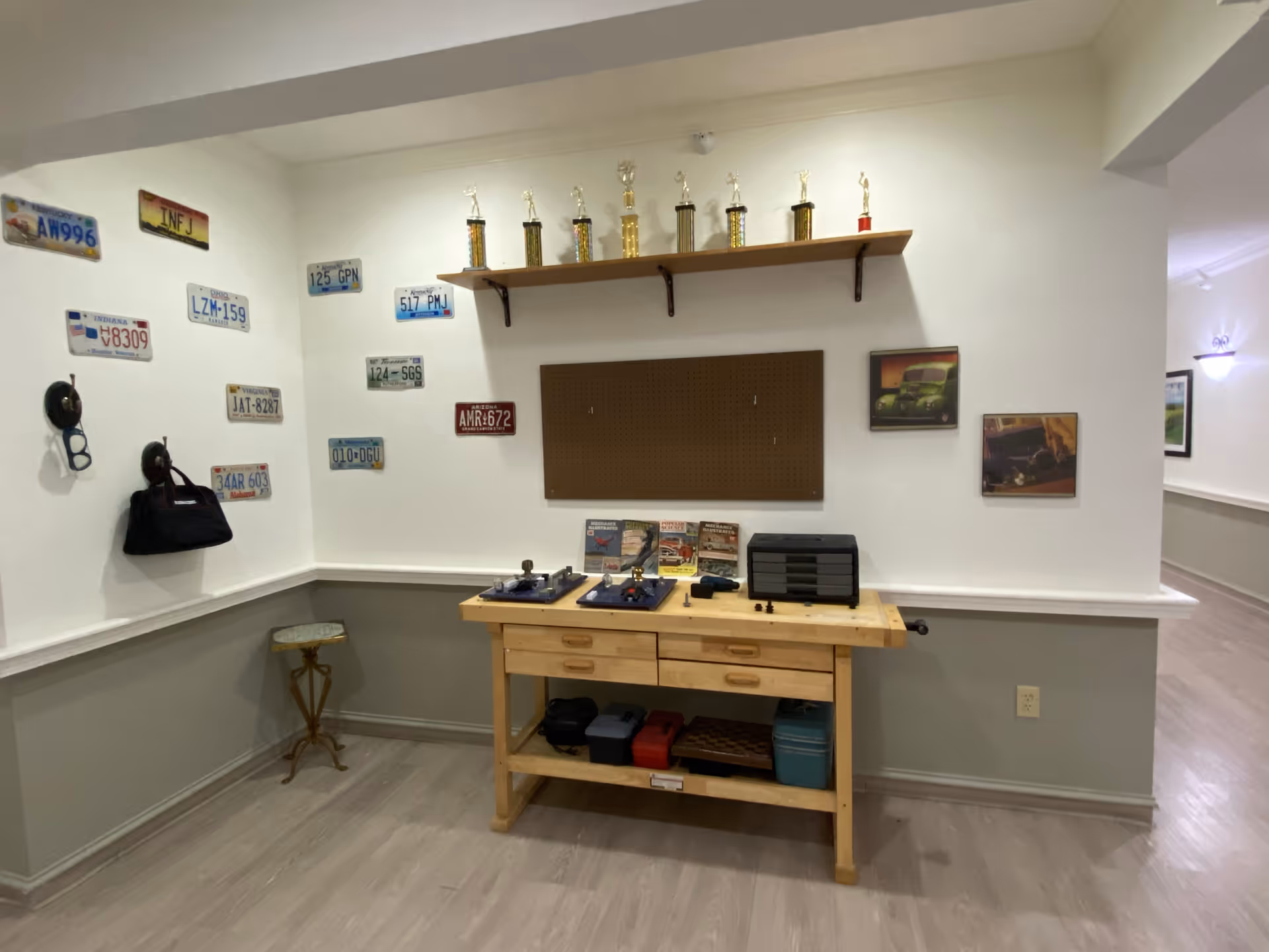 A small interior workshop corner with a wooden workbench, tools and storage beneath, license plates and pictures on the walls, and a shelf of trophies above.