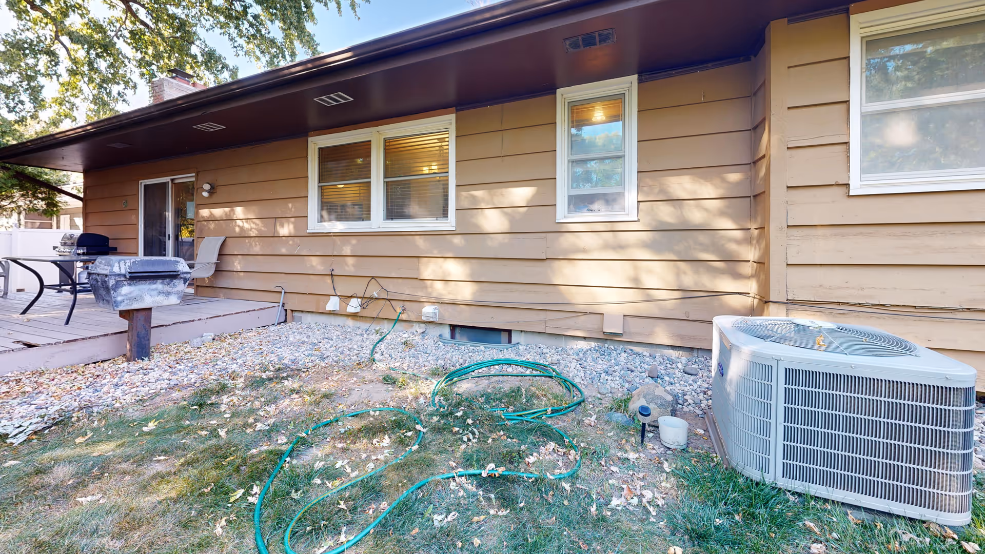 Back exterior of a single-story house with a small deck, patio furniture, an air conditioning unit, and garden hoses on the lawn.