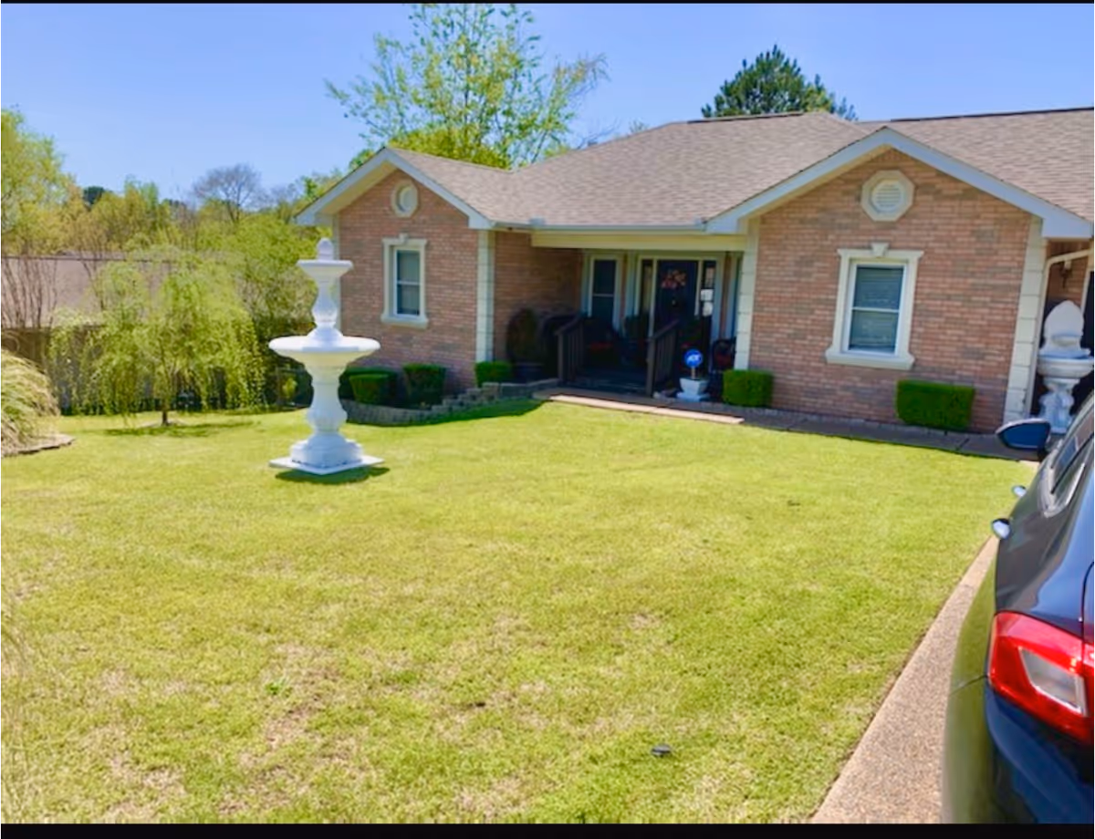 Front exterior view of a single-story brick house with a well-maintained lawn, a white decorative fountain in the yard, some bushes near the house, and a car parked on the driveway to the right.