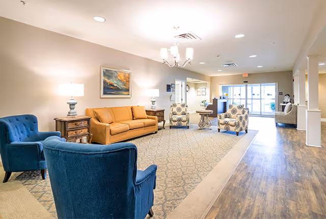 A spacious and well-lit living room area in a senior living facility named Wayman Place, featuring a mustard yellow sofa, two blue armchairs, two patterned armchairs, wooden side tables with lamps, a patterned area rug, and a painting on the wall. The room has wood flooring and large glass doors leading outside.