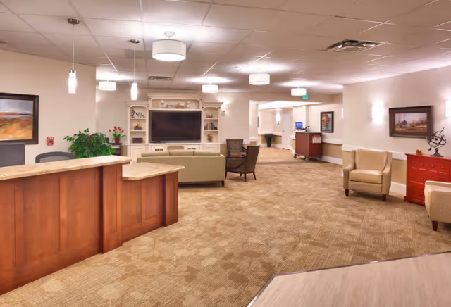 A spacious and well-lit senior living facility common area with a wooden reception desk on the left, beige carpeted floor, comfortable seating including sofas and armchairs, a large flat-screen TV mounted on a white entertainment unit, and decorative paintings and plants on the walls.