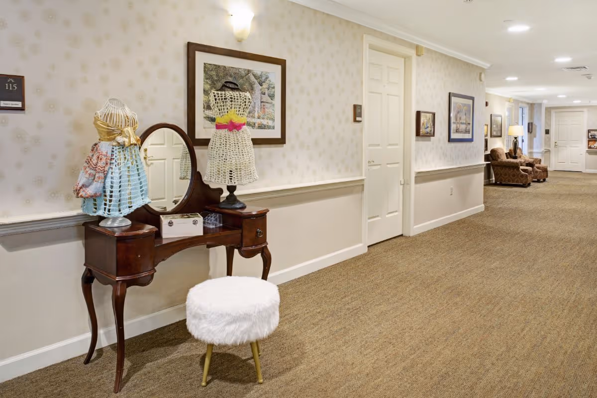Carpeted interior hallway with a vintage vanity table topped with decorative dress forms and a fluffy stool, leading to seating and doors down the corridor.