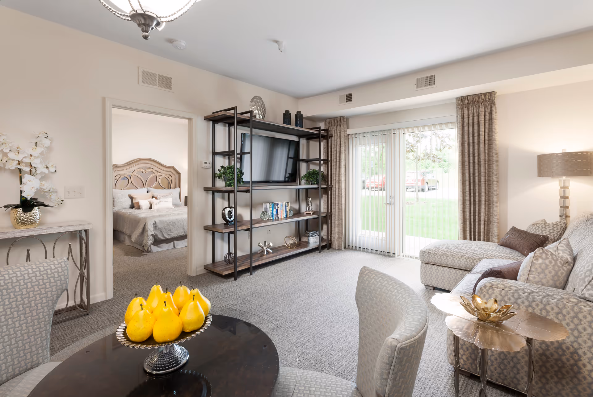 A bright living room with a patterned sofa and matching chair, a round table with a bowl of yellow pears, a metal and wood shelving unit with a TV and decorative items, and glass doors with vertical blinds leading outside. A bedroom with a bed and pillows is visible through an open doorway.