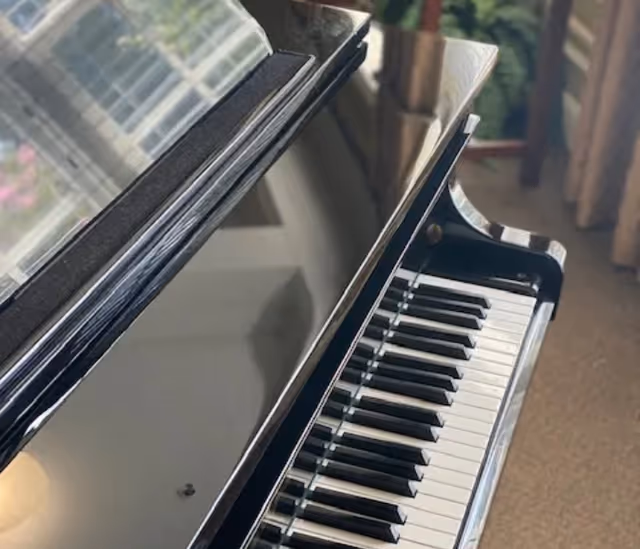Close-up of a glossy black grand piano keyboard and lid reflecting a window and curtains.