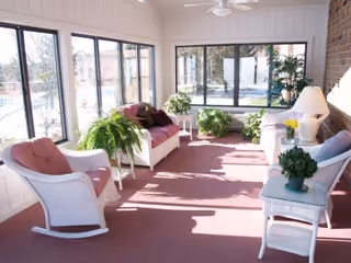 A bright sunroom with large windows on two sides, featuring white wicker furniture including chairs and sofas with pink cushions. Several green potted plants are placed around the room, and a table lamp is on a side table. The floor is covered with a pink carpet.