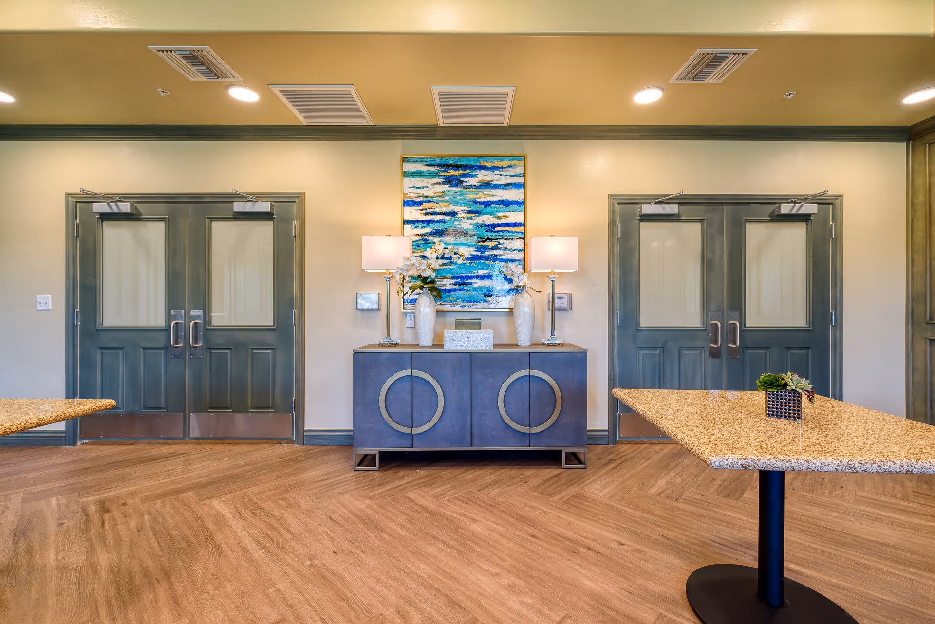 Interior view of a senior living facility room with two sets of double doors painted dark green. Between the doors is a blue sideboard with circular handles, topped with two white vases holding flowers and two lamps. Above the sideboard hangs a colorful abstract painting with blue, white, and yellow tones. The floor is wood with a herringbone pattern, and two granite tables with black pedestal bases are partially visible on either side of the image.