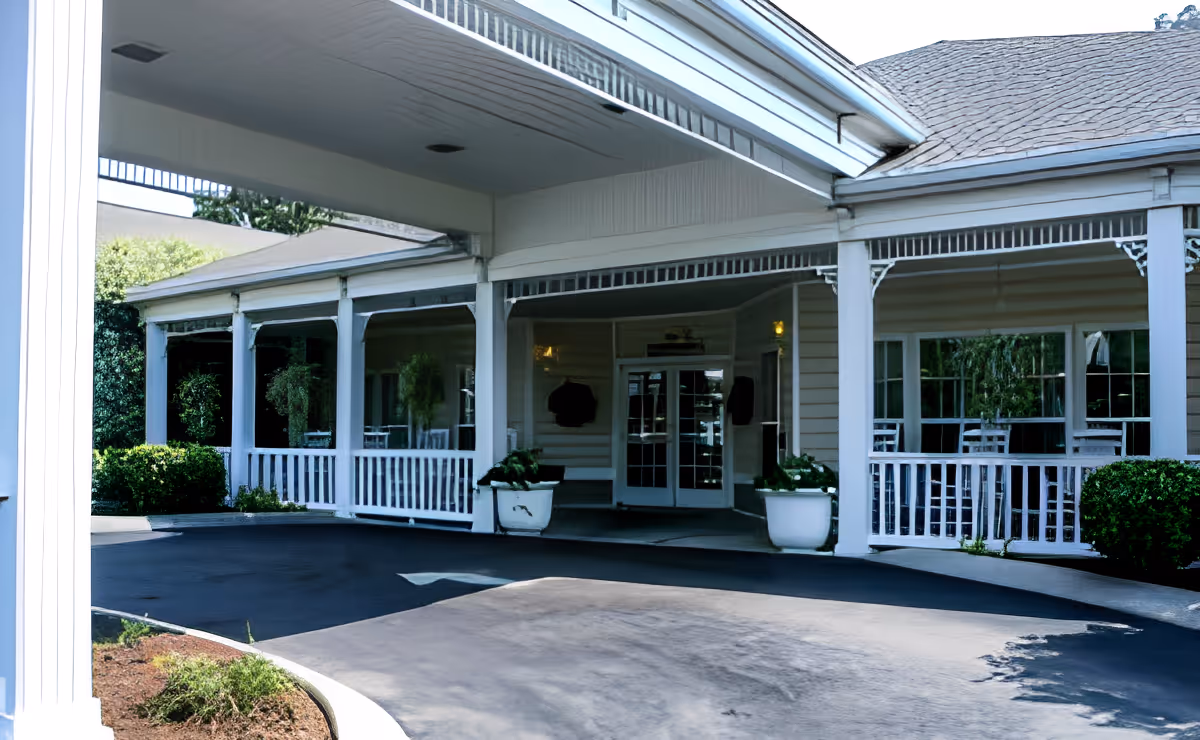 Covered front entrance and drive-through portico of a senior living facility with white columns, railings, potted plants and glass double doors.