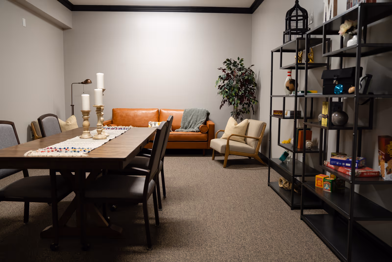 Cozy communal room with a wooden dining table and chairs, a leather sofa and armchair, and shelving with decorative items.