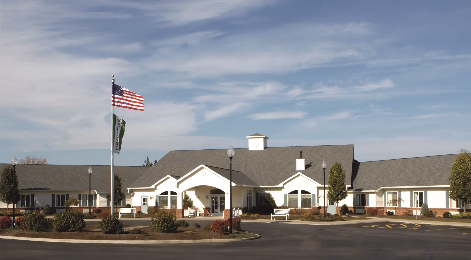 Exterior view of a single-story senior living facility building with a gray roof and white walls, surrounded by landscaped bushes and trees. There is an American flag and a welcome flag on flagpoles in front of the building, along with benches and street lamps near the entrance under a partly cloudy sky.