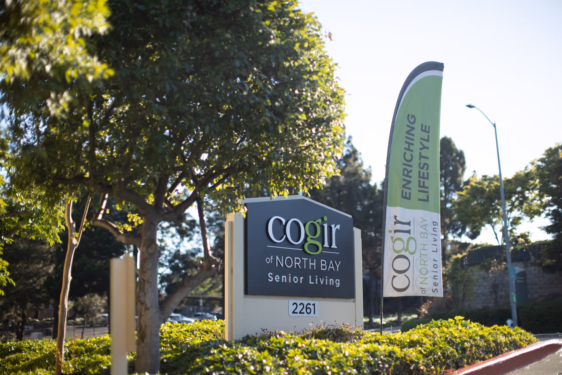 Outdoor view of the entrance sign for Cogir of North Bay Senior Living surrounded by greenery and trees, with a tall flag banner beside it displaying the facility's name and the phrase 'Enriching Lifestyle'.