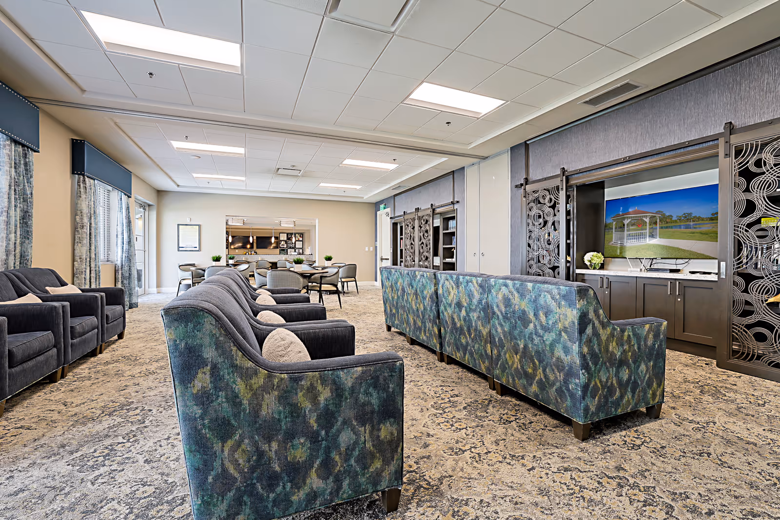 A spacious senior living common area with patterned blue and green sofas facing a large flat-screen TV mounted on a wall with decorative sliding panels. Additional seating includes dark armchairs and a dining table with chairs in the background near a kitchen pass-through window. The room has carpeted floors, large windows with curtains, and bright overhead lighting.