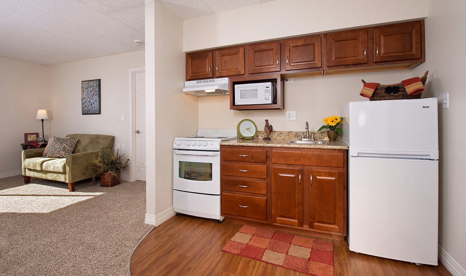A small kitchen area with wooden cabinets, a white stove, microwave, and refrigerator. There is a countertop with a sink, a decorative clock, a rooster figurine, and a vase with yellow flowers. To the left, there is a green loveseat with patterned cushions, a side table with a lamp, and a framed picture on the wall. The floor transitions from wood in the kitchen area to carpet in the living space.