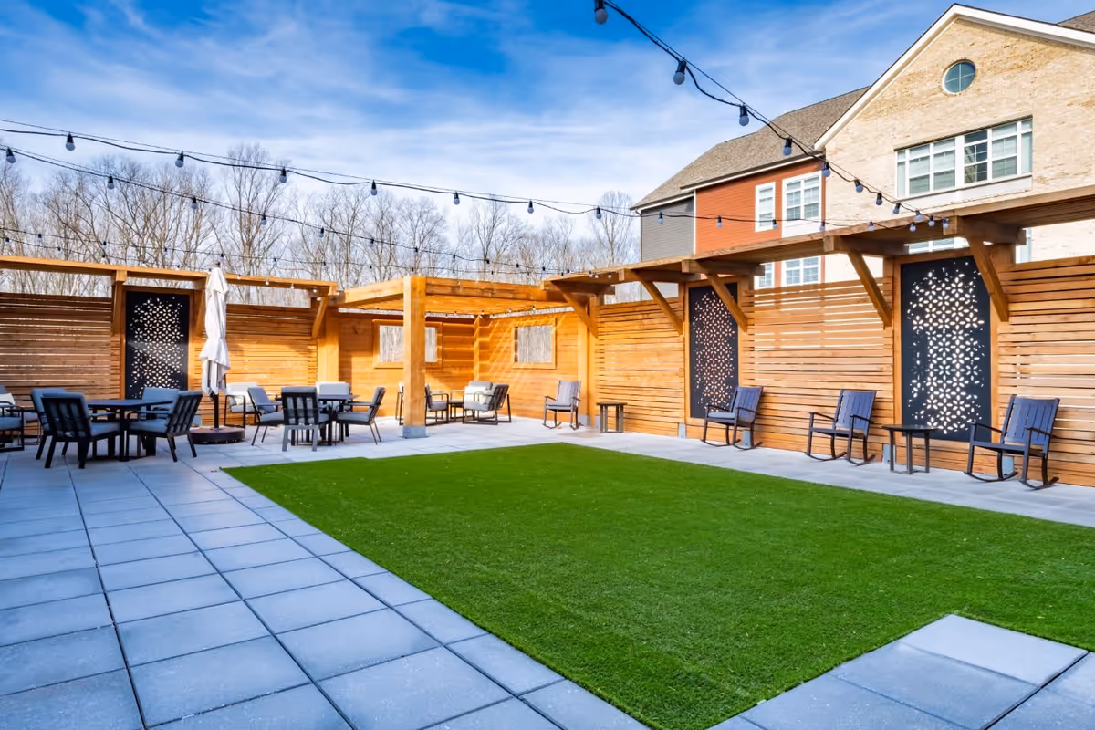 Outdoor patio area with artificial grass in the center, surrounded by gray tiled flooring. There are multiple seating arrangements with black chairs and small tables along wooden privacy walls. String lights are hung above the space, and a building with windows is visible in the background under a blue sky.