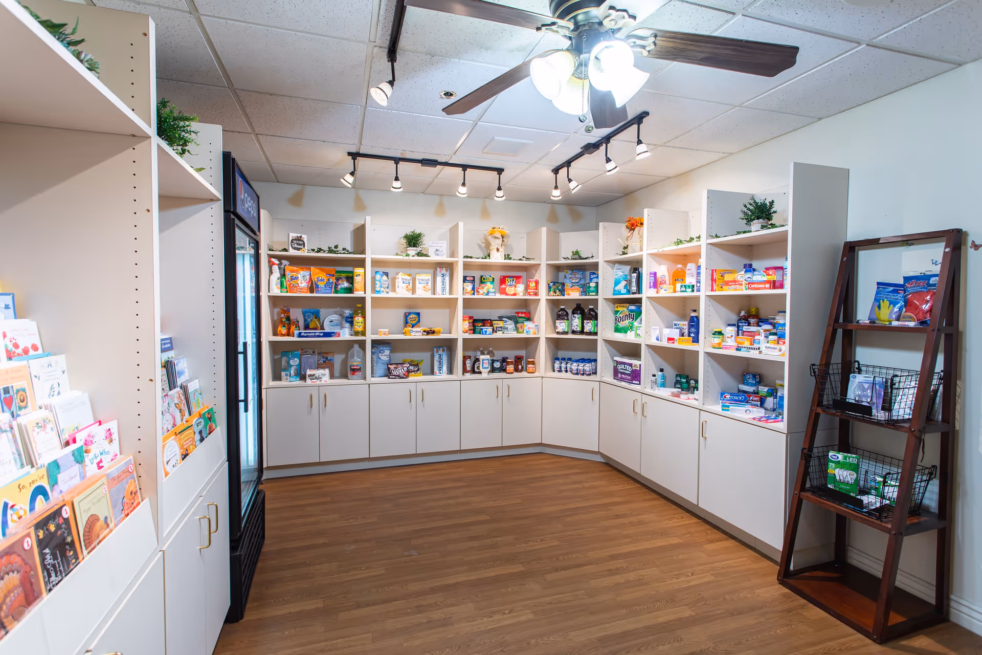 Small interior shop/pantry with white shelving stocked with snacks, household items, greeting cards, and a refrigerated display.