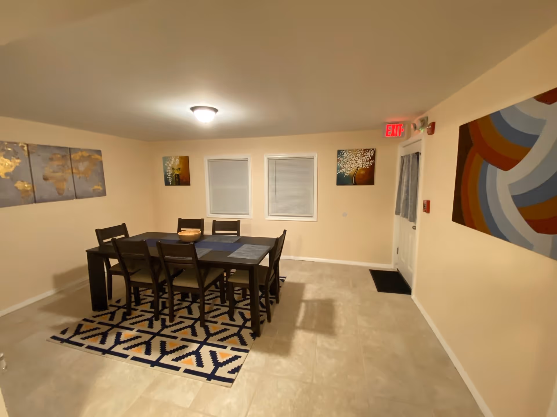 A dining room with a rectangular dark wooden table and six matching chairs placed on a patterned rug. The walls are light beige and decorated with three paintings: one large abstract piece on the right wall, a triptych world map on the left wall, and two smaller floral paintings between two windows on the back wall. There is a ceiling light fixture and an exit door with a red exit sign above it.