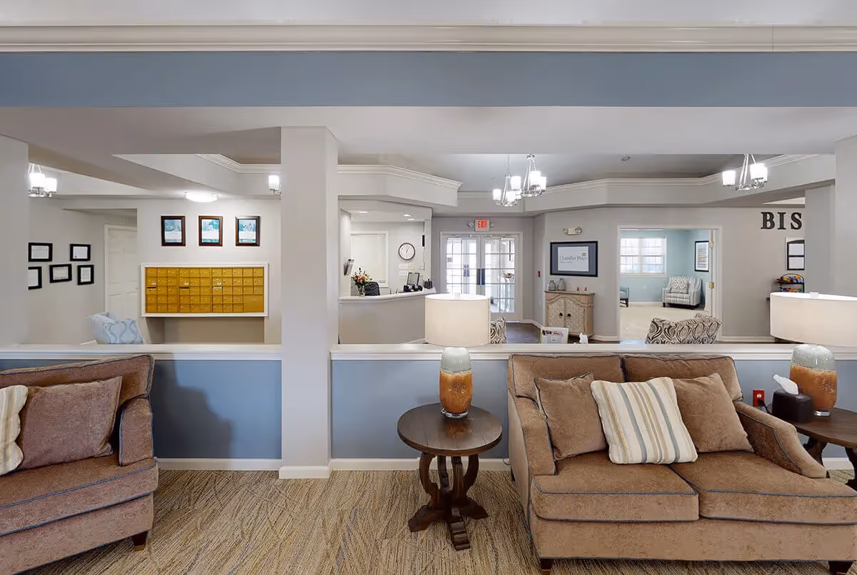 A cozy senior living facility lobby area with beige couches and striped pillows, a wooden side table with a lamp, and a reception desk in the background. The walls are painted in light blue and white, and there are framed pictures and mailboxes on the walls. A doorway leads to another room with additional seating.
