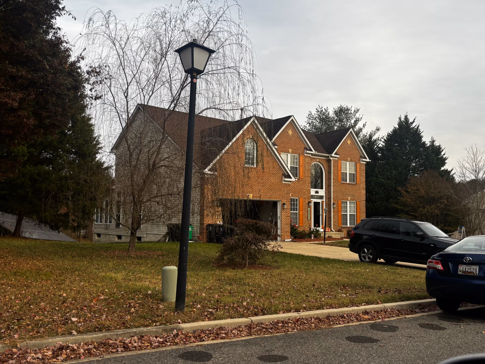 A two-story brick house with white trim and orange shutters, a driveway with two parked cars, a leaf-covered lawn, a street lamp, and several trees including a bare tree in front of the house under a cloudy sky.