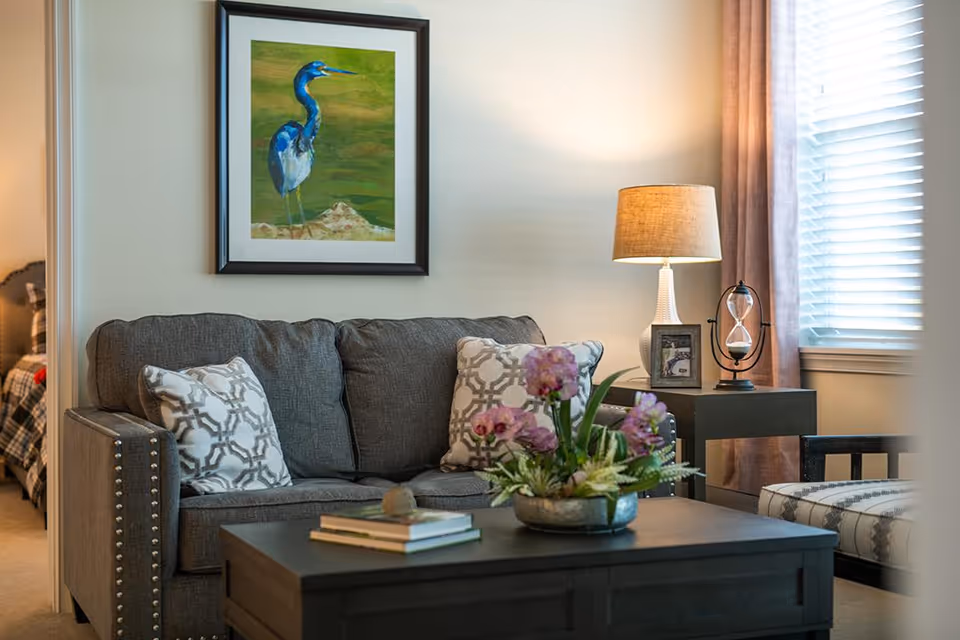 A cozy living room with a gray sofa adorned with patterned pillows, a dark wooden coffee table with books and a flower arrangement, a side table with a lamp, a framed photo, and an hourglass. A window with blinds and curtains lets in natural light, and a framed painting of a blue heron hangs on the wall above the sofa. A glimpse of a bedroom with a plaid bedspread is visible through an open doorway.