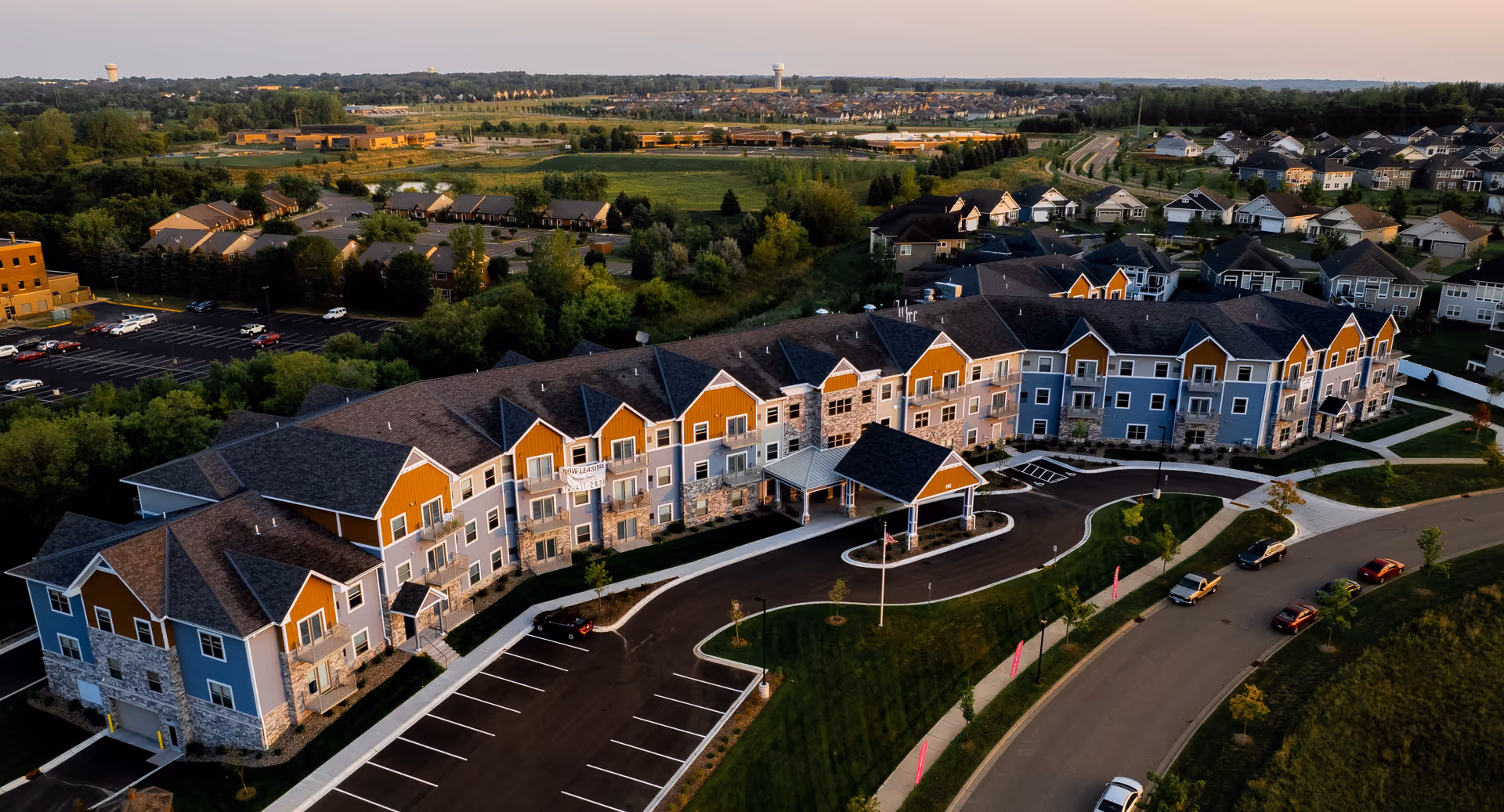 Aerial view of a large multi-story senior living building with driveways, parking lots, and surrounding homes at sunset.