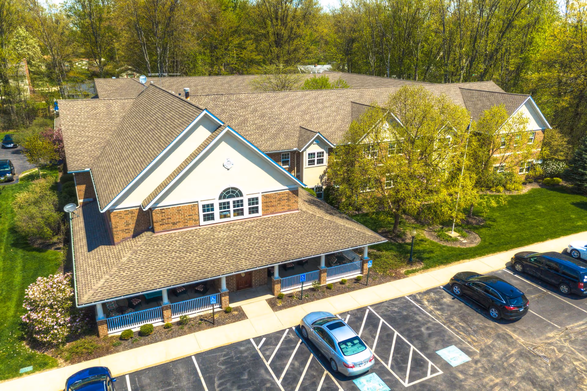 Aerial view of Symphony at Olmsted Falls senior living facility showing a large brick building with a gabled roof, surrounded by trees and greenery. There is a parking lot in front with several cars parked, including designated handicap parking spaces. The building has a covered porch area with seating.