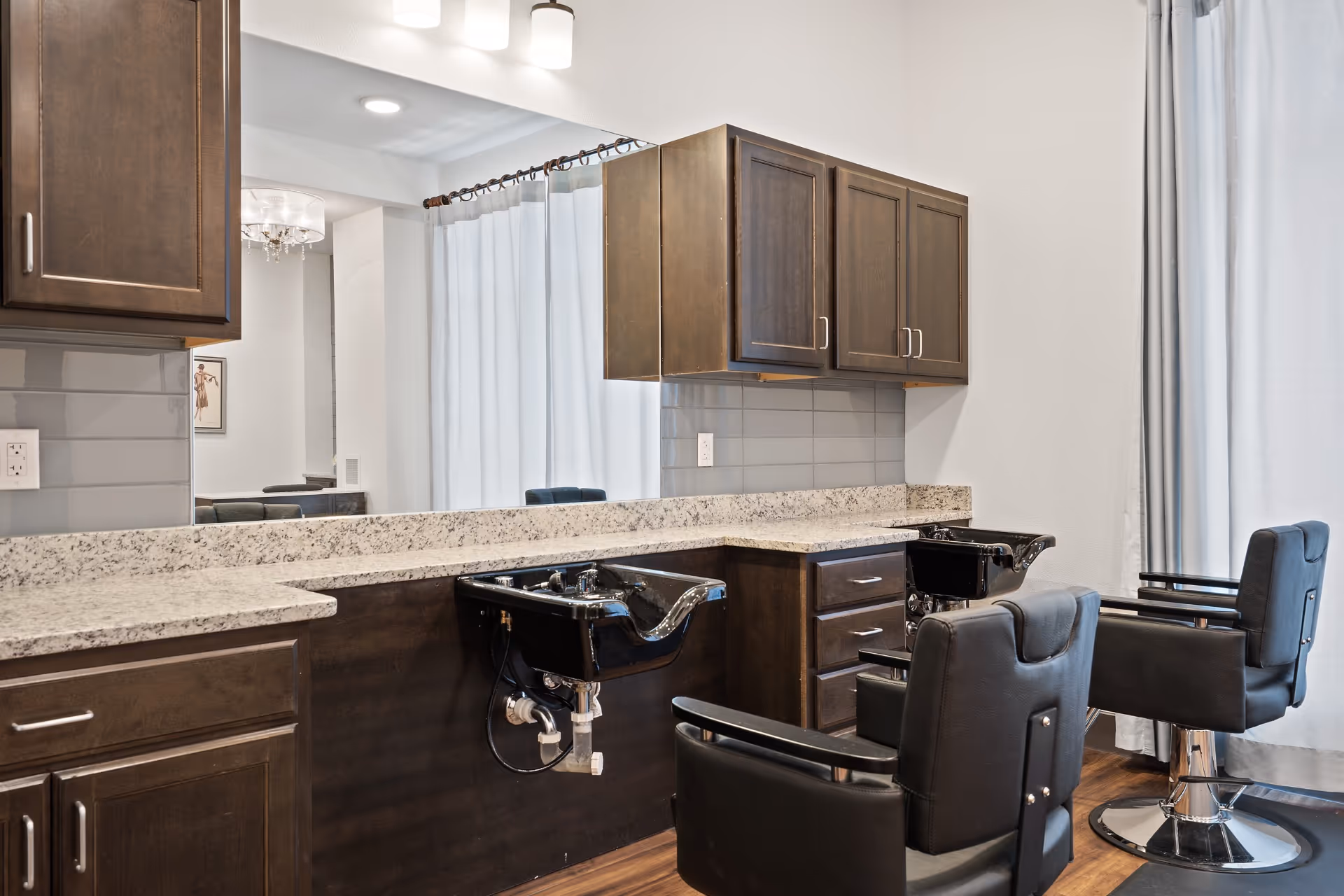 Interior view of a salon area with two black salon chairs and two black hair washing sinks mounted on a dark wood cabinet with granite countertops. A large mirror is mounted on the wall above the sinks, and there are upper cabinets for storage. The floor is wooden, and light curtains cover the windows.