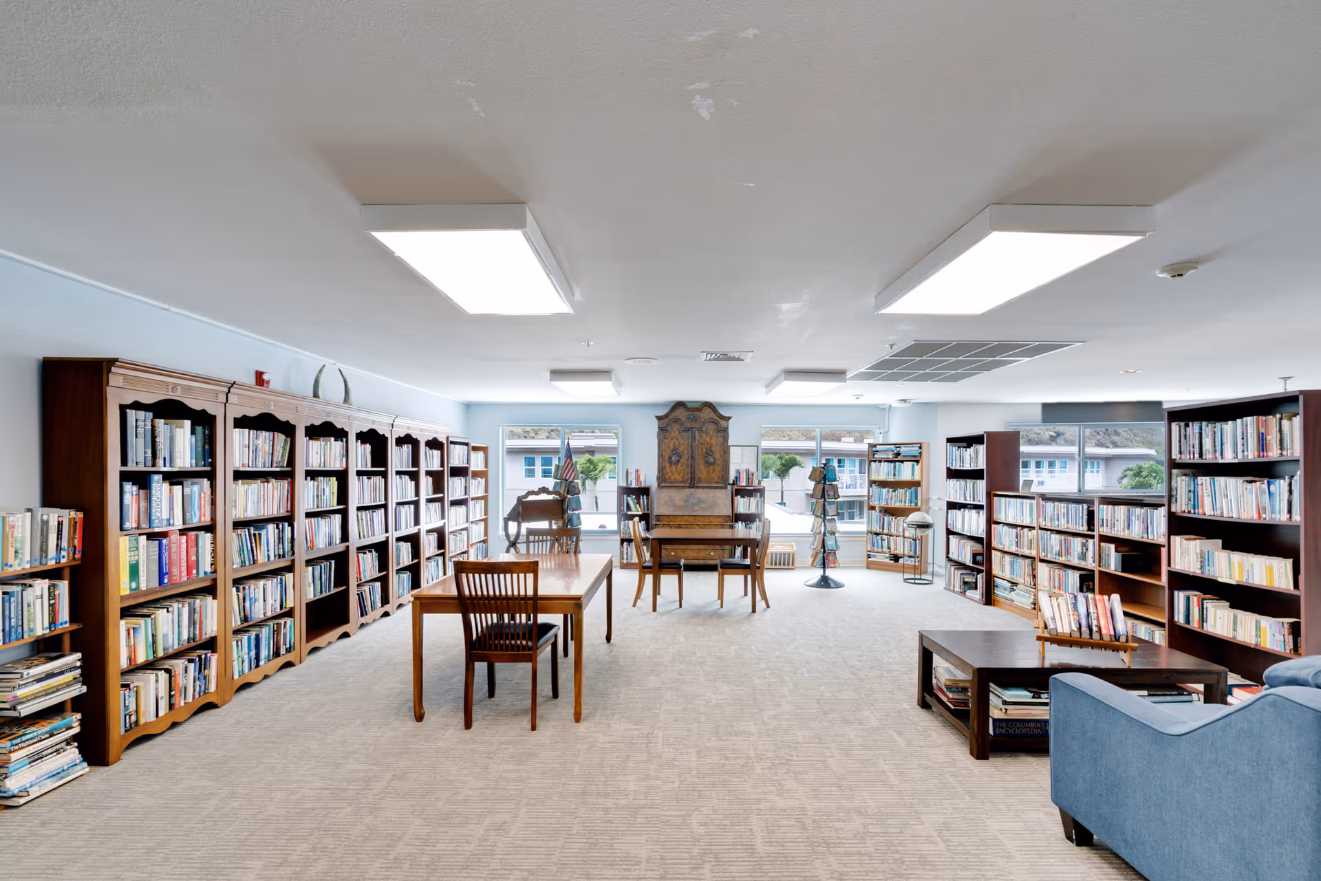 A spacious library room with multiple wooden bookshelves filled with books along the walls. There are two wooden tables with chairs in the center of the room, a blue armchair, and a coffee table with books on it. Large windows at the far end let in natural light, and the room has a light-colored carpet and ceiling lights.
