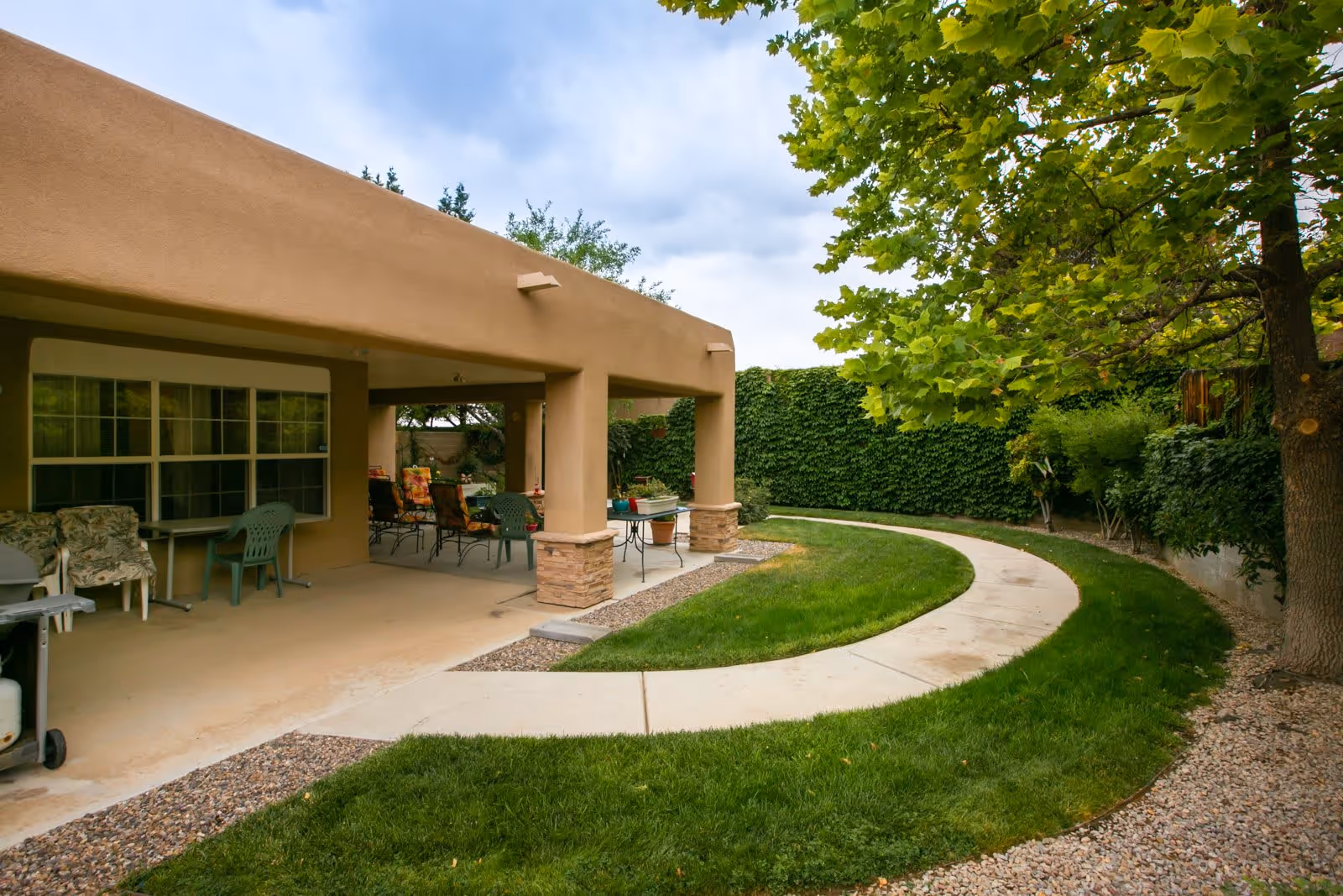 Outdoor patio area with a covered seating space featuring chairs and tables. The patio is adjacent to a curved concrete pathway surrounded by green grass, trees, and a tall hedge providing privacy.