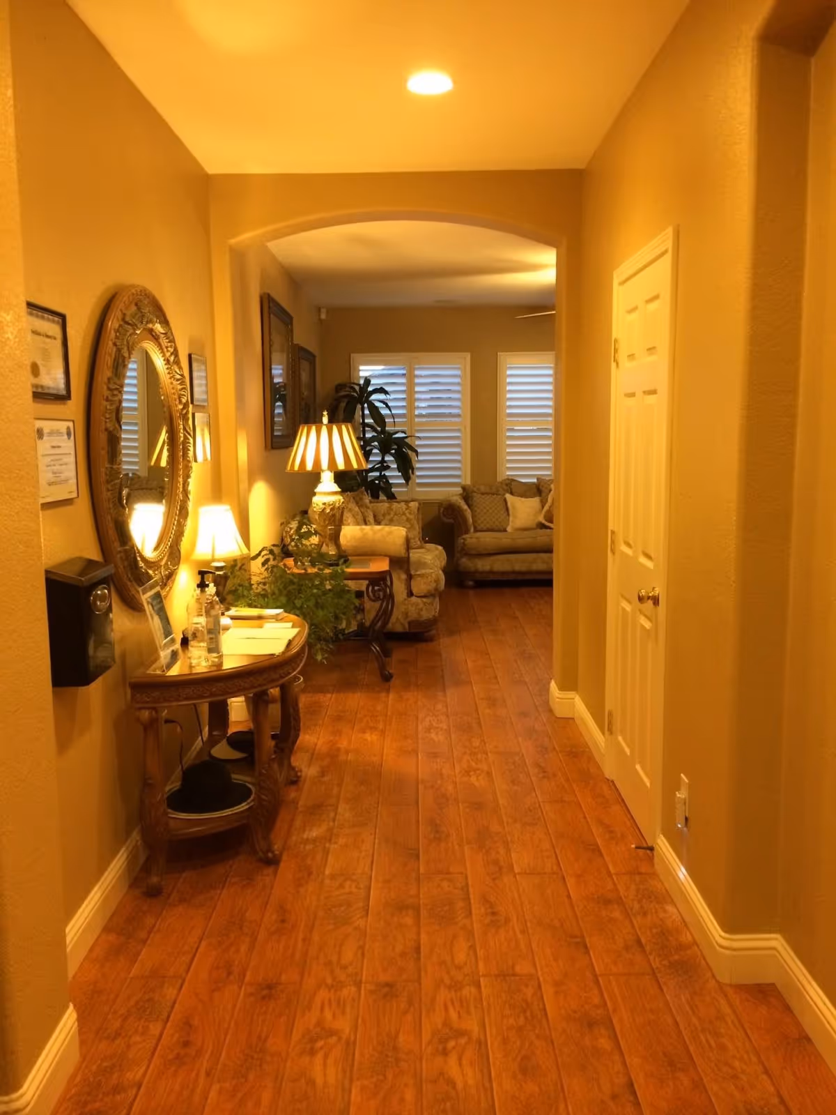 A warmly lit hallway with wooden flooring leading to a living room area. On the left side of the hallway, there is an ornate round mirror above a wooden console table with lamps, plants, and various items. The living room at the end has cushioned sofas, a side table with a lamp, and windows with white shutters.