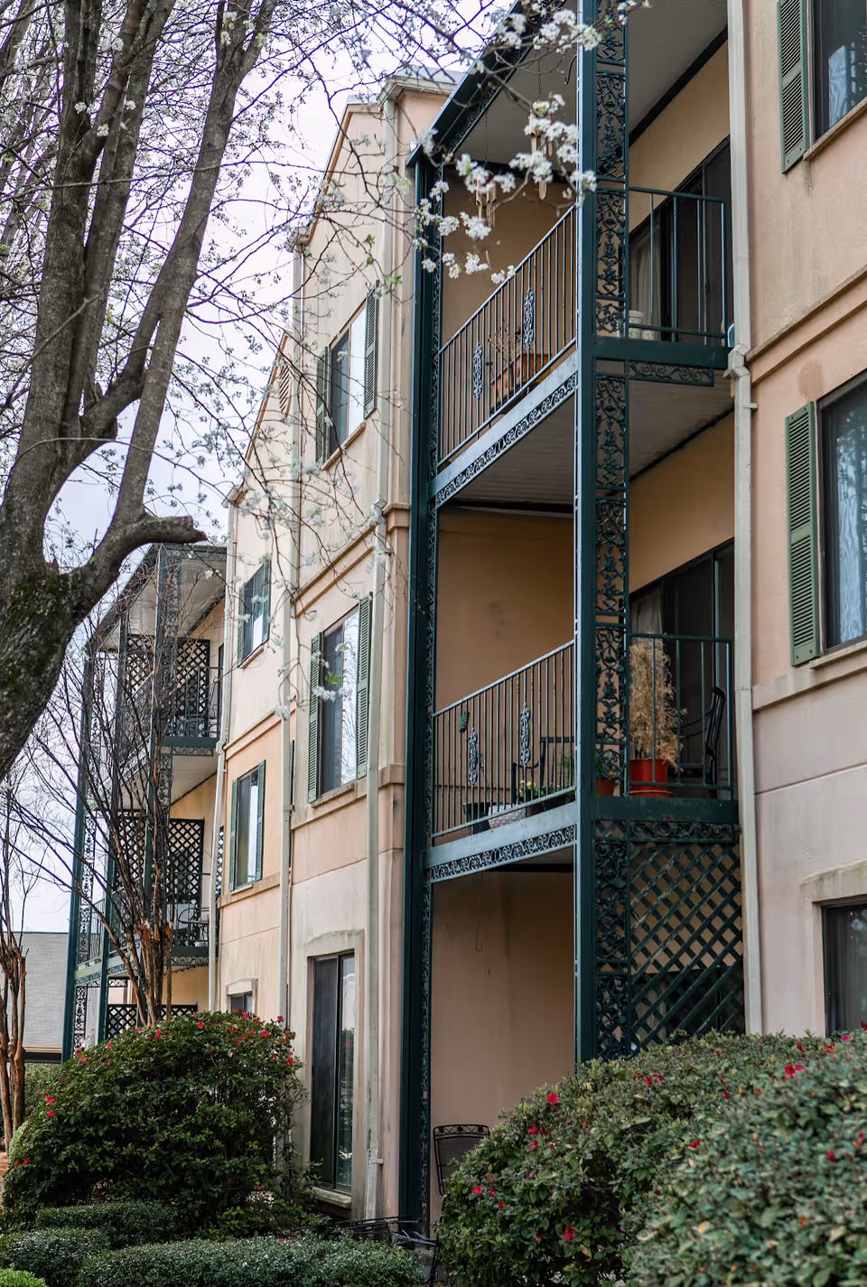 Exterior view of a multi-story residential building with beige walls and green metal balconies. There are trees with budding flowers and well-maintained bushes in the foreground.