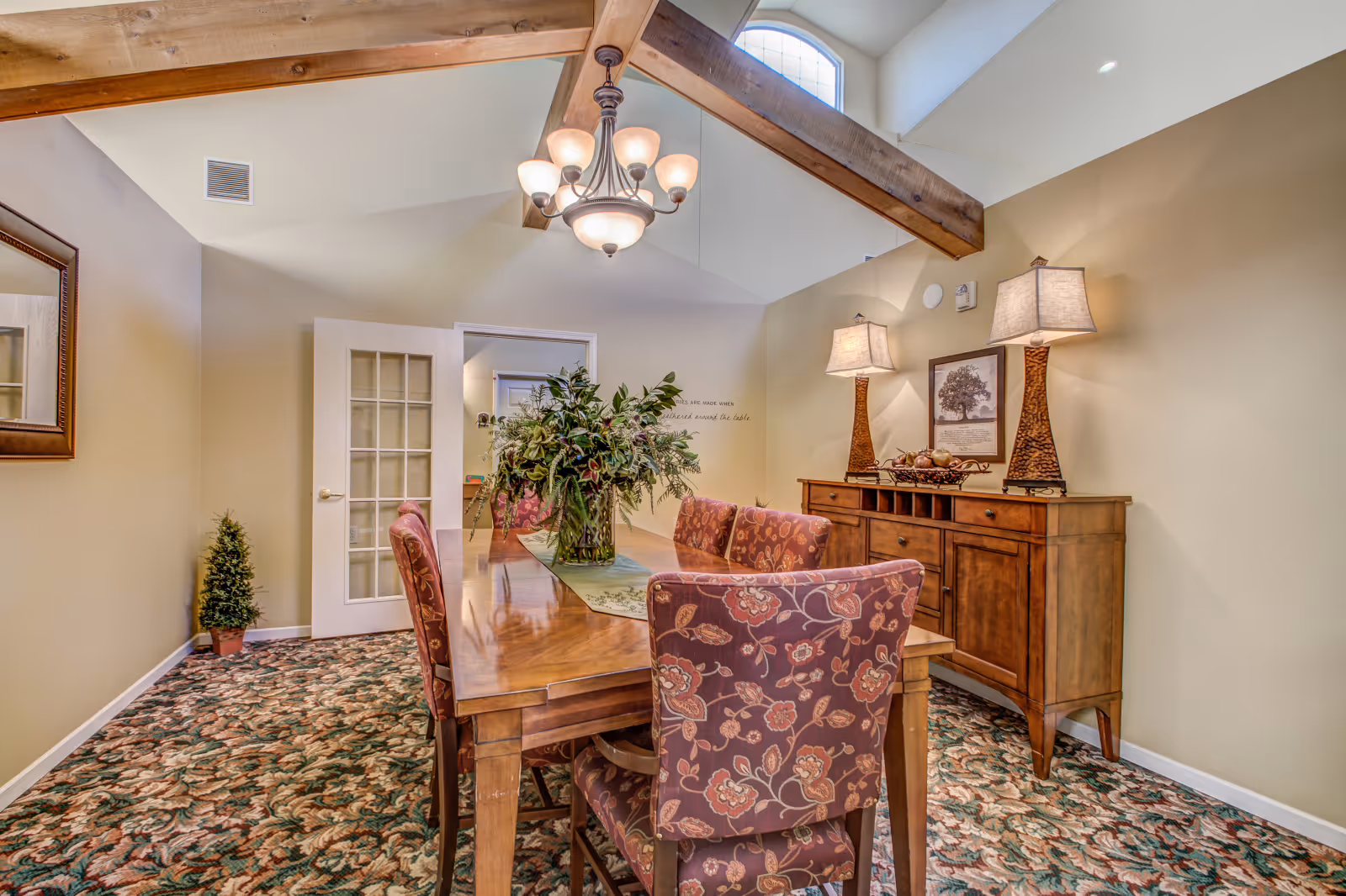 A dining room with a wooden table surrounded by floral upholstered chairs. A large floral centerpiece sits on the table. The room has a patterned carpet, beige walls, and a wooden sideboard with two lamps and a framed picture above it. Exposed wooden beams and a chandelier hang from the vaulted ceiling with a small arched window.