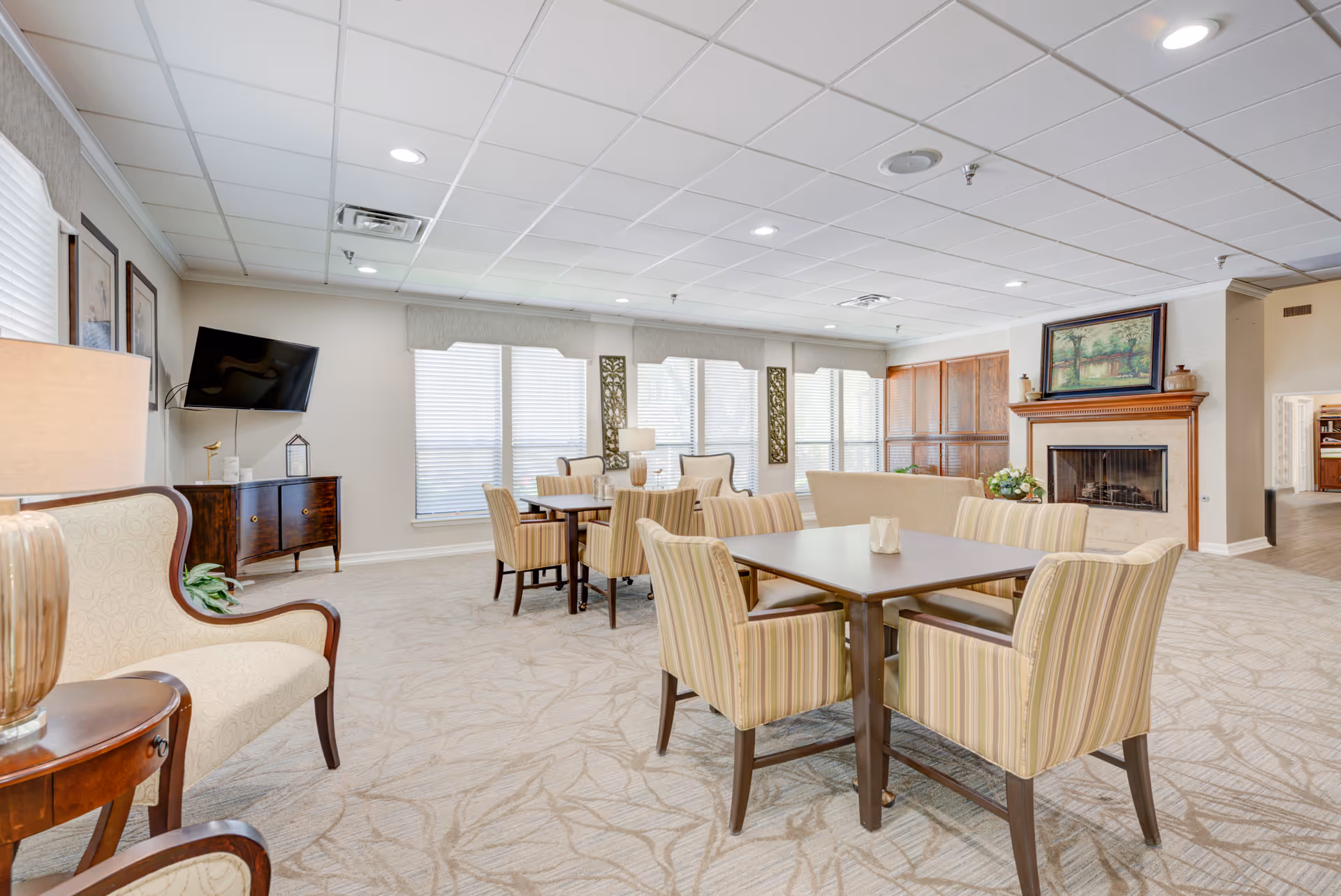 A bright and spacious common area in a retirement community featuring multiple tables with striped upholstered chairs, a fireplace with a painting above it, a wall-mounted TV, and large windows with blinds allowing natural light to fill the room.