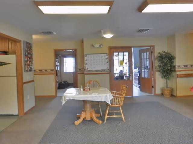 Interior view of a room with a small wooden table covered with a white tablecloth and two wooden chairs. The room has beige walls with a decorative border and carpeted floor. There are two doorways in the background, one with a wooden door and glass panels, and the other an open doorway. A potted plant is placed near the right wall, and a calendar is hanging on the wall between the doorways.