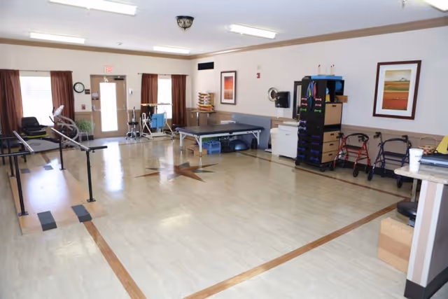 A spacious rehabilitation room with exercise equipment including parallel bars, walkers, a therapy table, and storage shelves with various therapy supplies. The room has light-colored flooring with a star design in the center, beige walls, framed artwork, and large windows with brown curtains allowing natural light.