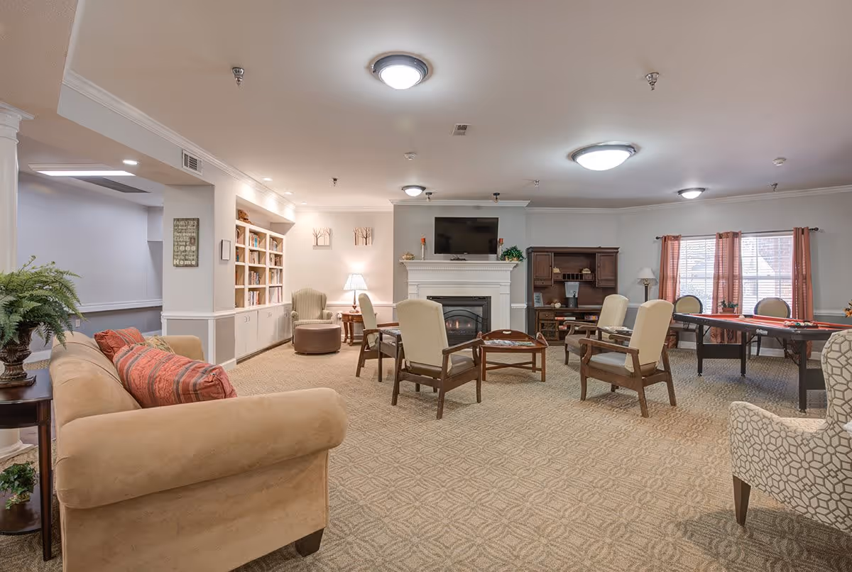 A spacious senior living community lounge with beige carpet and neutral walls. The room features a beige sofa with red patterned pillows, several armchairs arranged around a coffee table, a fireplace with a TV mounted above it, a bookshelf filled with books, a wooden cabinet, and a table near windows with red curtains. The room is well-lit with ceiling lights and a table lamp.